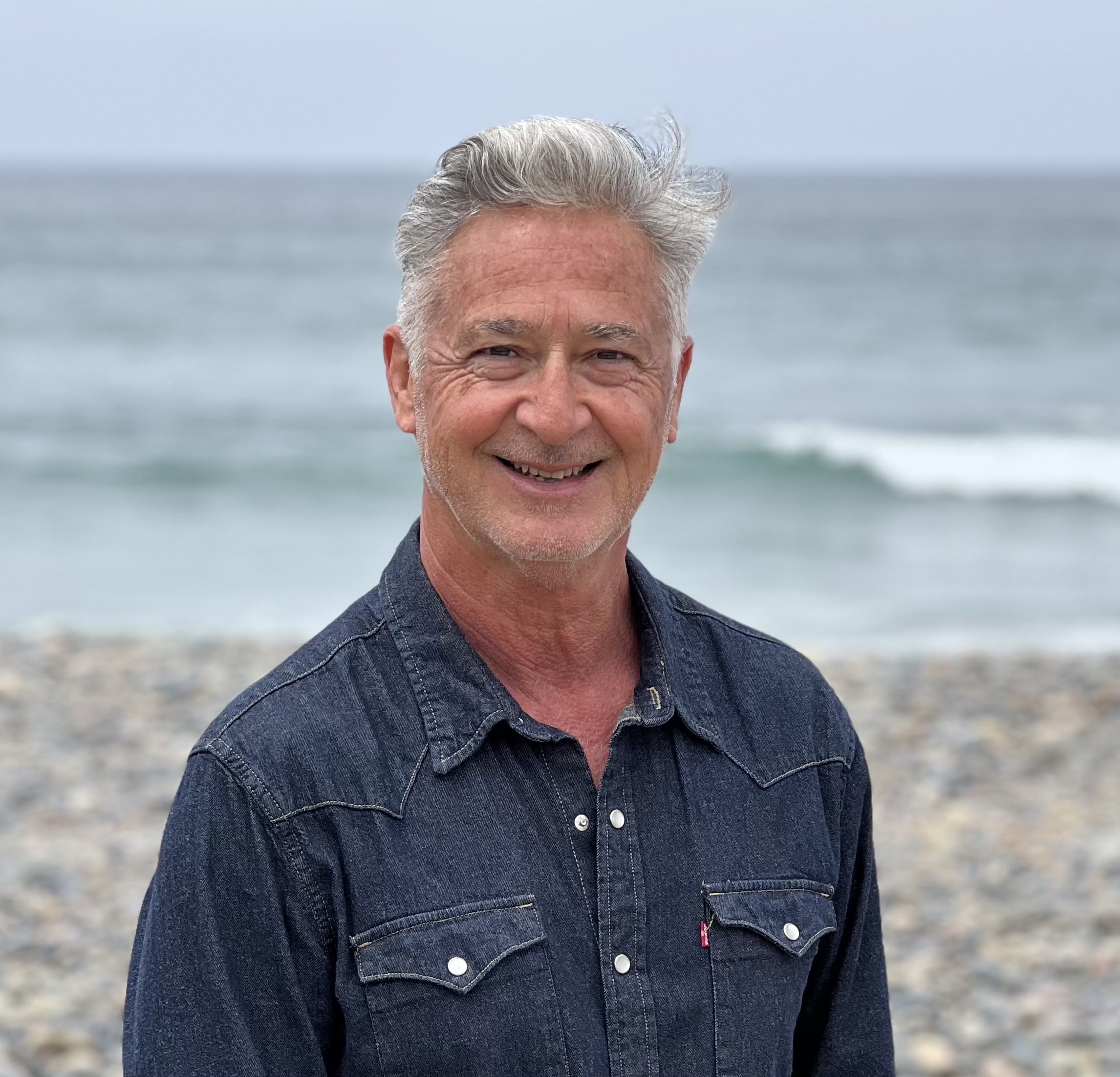 A man with grey hair and a blue button up smiles at the camera on the beach.