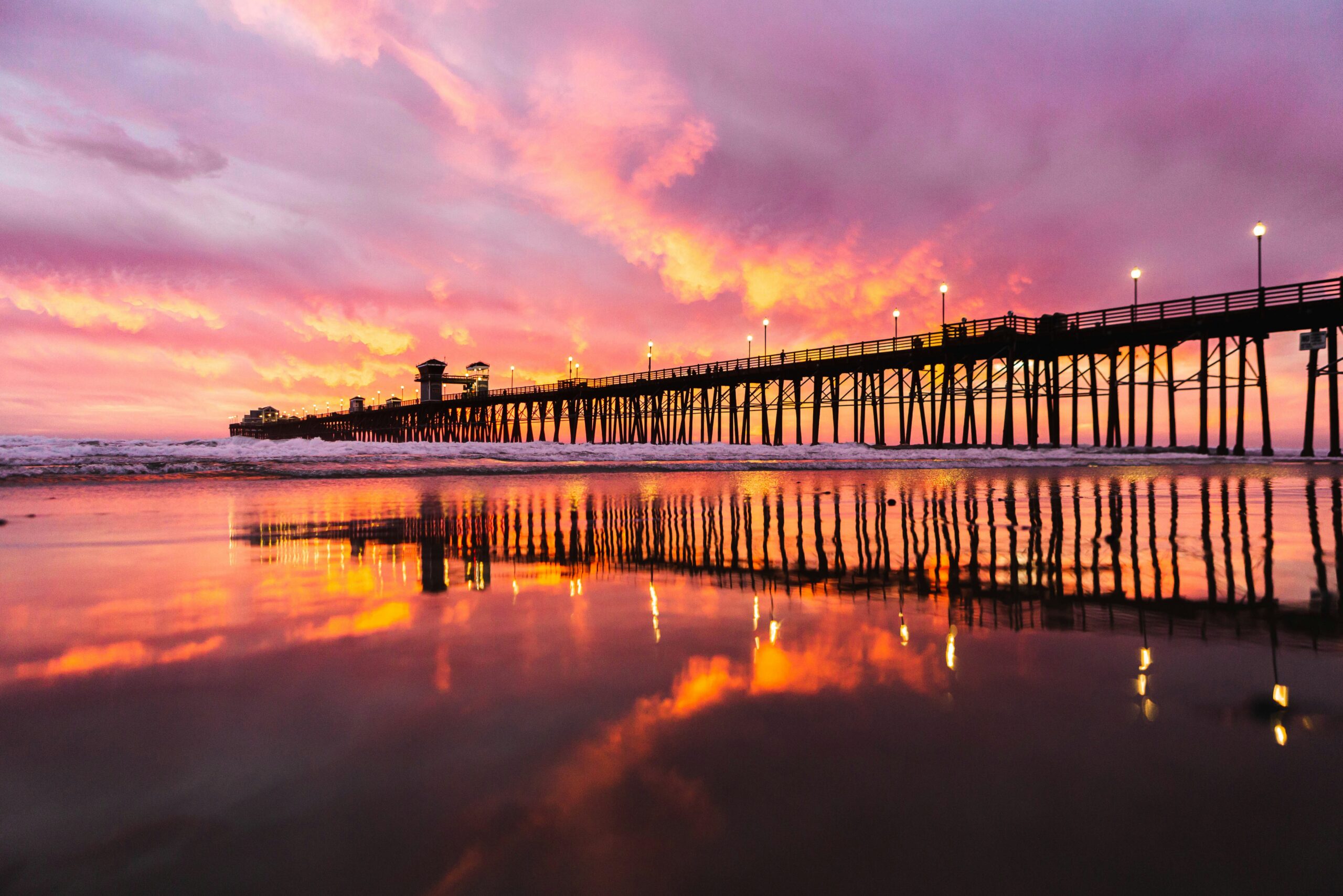45-degree view of the Oceanside Pier with a pink, purple, and gold sunset behind it.